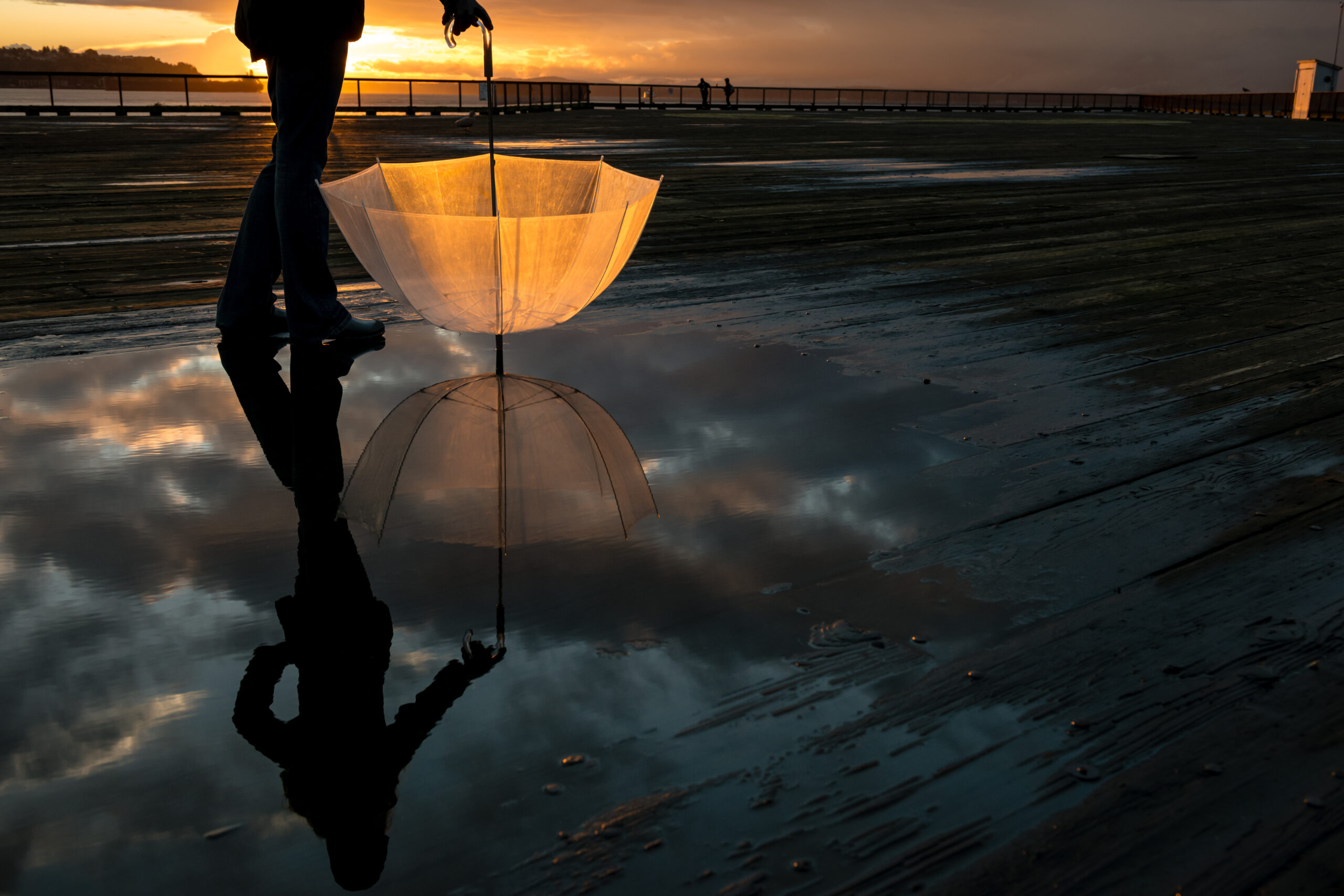 Eine Frau hält einen durchsichtigen Regenschirm, der Himmel ist stürmisch und leuchtet im orange des Sonnenuntergangs.