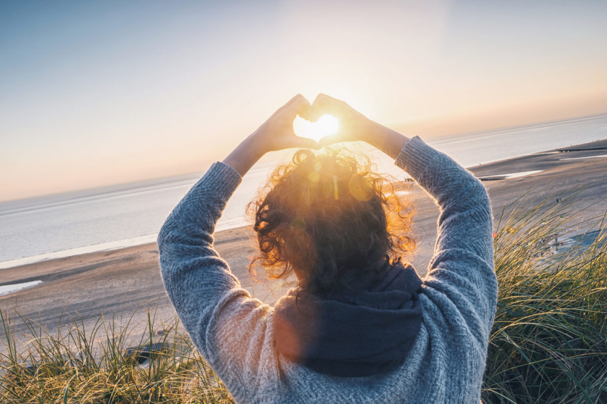 Eine Frau steht am Strand und formt beide Hände als Herz über ihrem Kopf, dahinter leuchtet der Sonnenuntergang
